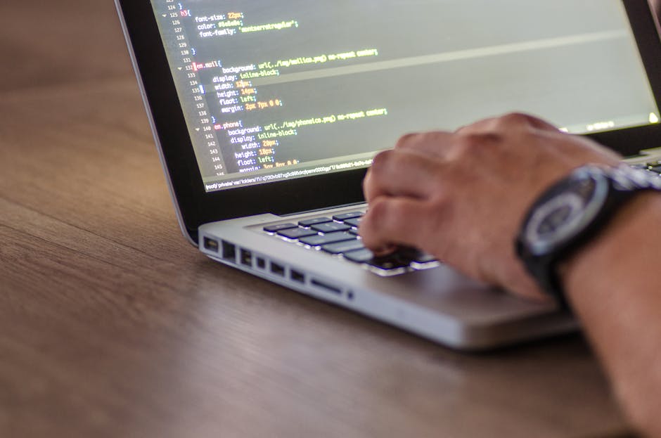 A close-up image of a person's hand wearing a black wristwatch, typing on a silver laptop placed on a wooden surface. The laptop screen displays lines of code with colorful syntax highlighting, indica